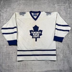 May include: White hockey jersey with blue detailing. The jersey has a V-neck and features the text "TORONTO MAPLE LEAFS" within a blue maple leaf logo. Blue stripes are on the sleeves and hem.