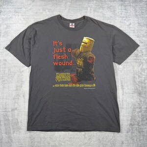 May include: A charcoal gray short-sleeve t-shirt featuring a graphic of a knight in a helmet with missing arms. The text reads: It's Just a flesh wound... MONTY PYTHON ...come back here and I'll bite your kneecaps off! The graphic includes red blood.