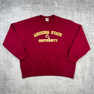 Può includere: Una felpa bordeaux con la scritta "ARIZONA STATE UNIVERSITY" in giallo. La felpa ha un girocollo e maniche lunghe. Una grafica della mascotte bordeaux e oro è tra le parole "STATE" e "UNIVERSITY".