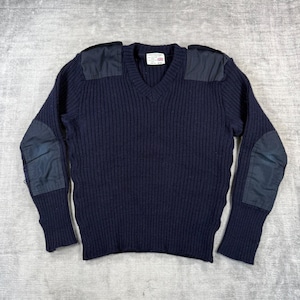 Op de afbeelding: Een donkerblauwe V-hals trui met een geribbelde breistructuur. Het kledingstuk is voorzien van effen donkerblauwe stoffen patches op beide schouders en aan de buitenkant van elke elleboog. Een wit rechthoekig label is in de hals genaaid.