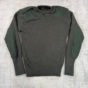 Op de afbeelding: Een donker olijfgroene trui van geribbeld breiwerk met een ronde hals, lange mouwen en effen donker olijfgroene stoffen patches op de schouders en ellebogen. De trui heeft een dik, gestructureerd verticaal ribpatroon over het hele kledingstuk.