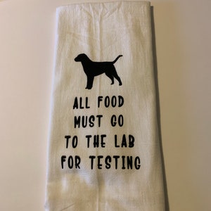 May include: White kitchen towel with a black silhouette of a dog and the text "ALL FOOD MUST GO TO THE LAB FOR TESTING". The towel is folded and has a slightly wrinkled texture.