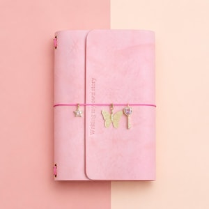 May include: A light pink journal with a vertical flap and the text Writing my own story embossed on the front. A bright pink elastic band wraps around the cover, featuring three gold-toned charms: a star, a butterfly, and a heart-topped key.