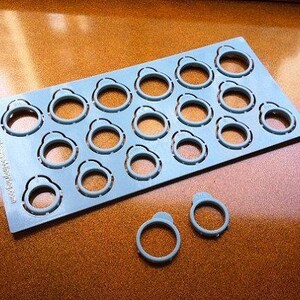 Pu&ograve; includere: Una piastra in plastica azzurra con una griglia di quindici misuratori di anelli circolari, con due misuratori aggiuntivi staccati e posizionati sotto. Il testo www.myjewelweddingring.com &egrave; stampato verticalmente lungo il bordo sinistro.