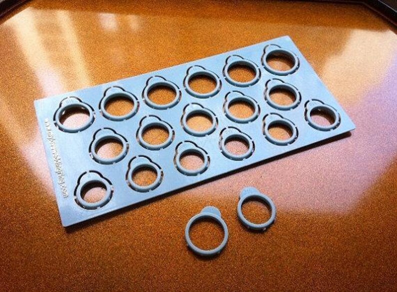 Pu&ograve; includere: Una piastra in plastica azzurra con una griglia di quindici misuratori di anelli circolari, con due misuratori aggiuntivi staccati e posizionati sotto. Il testo www.myjewelweddingring.com &egrave; stampato verticalmente lungo il bordo sinistro.