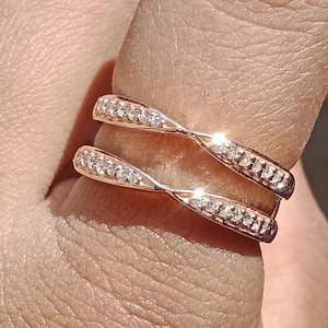 May include: Rose gold ring guard with two bands, each adorned with small, sparkling clear stones. The bands curve and cross over each other, creating a unique and elegant design. The ring is shown on a finger.