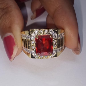 May include: A gold-toned ring featuring a large, rectangular, red gemstone at its center. The ring is adorned with smaller, clear gemstones and decorative gold accents. The design is symmetrical and the ring is held by a hand.