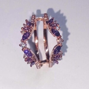 May include: Rose gold ring guard with a halo of purple and clear gemstones. The ring guard features marquise-cut purple stones and smaller clear stones, creating a sparkling, decorative design. The ring guard is designed to enhance the look of a solitaire ring.