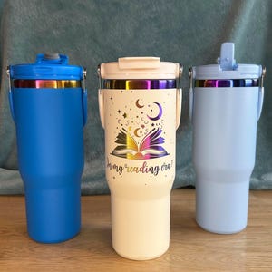 Puede incluir: Tres vasos aislados en azul, blanco roto y azul claro. El vaso blanco roto presenta un dise&ntilde;o de libro, estrellas y lunas con el texto "in my reading era!". Cada vaso tiene un asa y una tapa con una banda iridiscente met&aacute;lica.