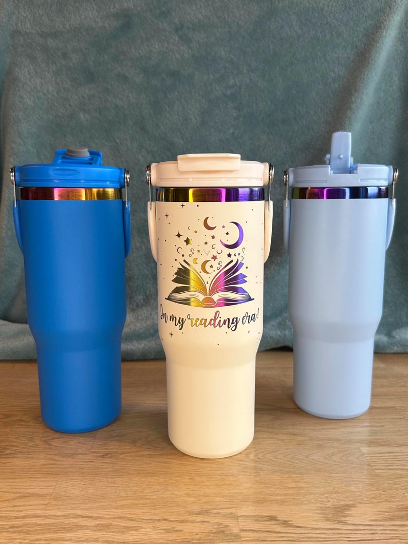 Puede incluir: Tres vasos aislados en azul, blanco roto y azul claro. El vaso blanco roto presenta un dise&ntilde;o de libro, estrellas y lunas con el texto "in my reading era!". Cada vaso tiene un asa y una tapa con una banda iridiscente met&aacute;lica.