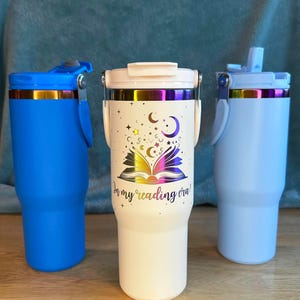 Puede incluir: Tres vasos aislados en azul, blanco y crema. El vaso crema presenta un dise&ntilde;o de libro abierto con estrellas, lunas y el texto "In my reading era!". Cada vaso tiene un asa y una tapa.