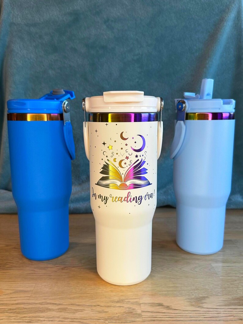 Puede incluir: Tres vasos aislados en azul, blanco y crema. El vaso crema presenta un dise&ntilde;o de libro abierto con estrellas, lunas y el texto "In my reading era!". Cada vaso tiene un asa y una tapa.