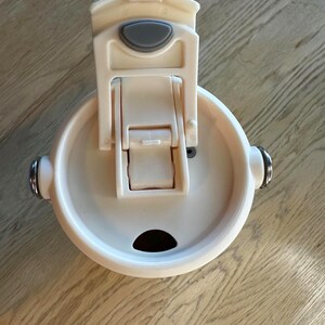 Puede incluir: Una tapa de taza de viaje de color crema con cierre abatible y un peque&ntilde;o bot&oacute;n de forma ovalada. La tapa tiene una abertura circular y dos detalles plateados en los laterales. La taza est&aacute; dise&ntilde;ada para mantener las bebidas calientes o fr&iacute;as.