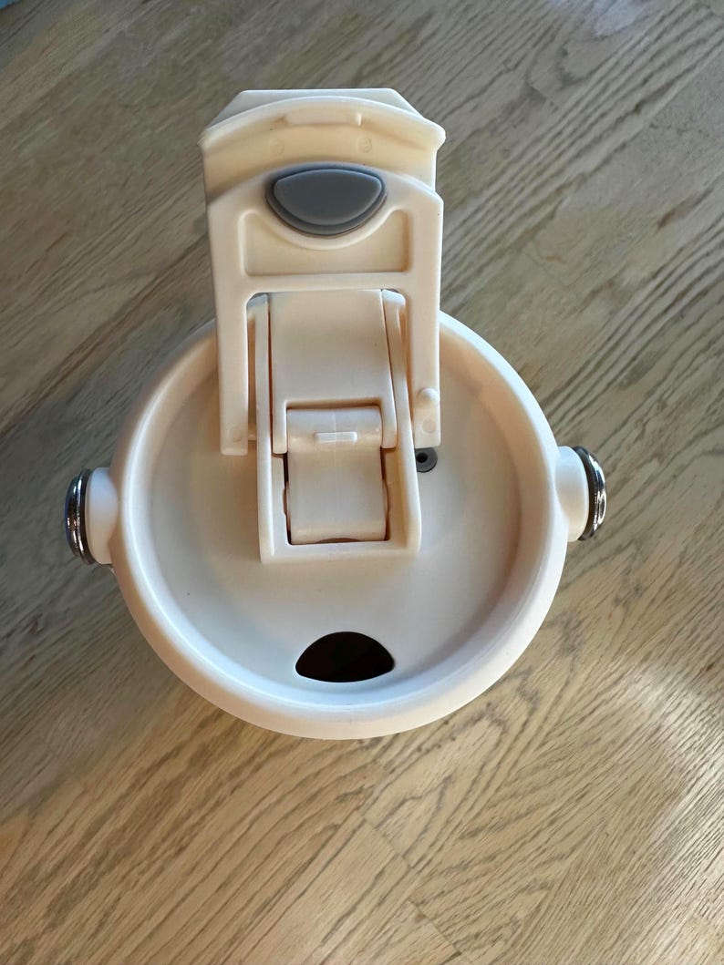 Puede incluir: Una tapa de taza de viaje de color crema con cierre abatible y un peque&ntilde;o bot&oacute;n de forma ovalada. La tapa tiene una abertura circular y dos detalles plateados en los laterales. La taza est&aacute; dise&ntilde;ada para mantener las bebidas calientes o fr&iacute;as.