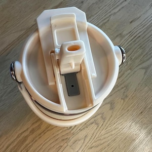 Puede incluir: Un termo de viaje color crema con tapa abatible y borde plateado. La tapa est&aacute; abierta, revelando la boquilla y un interior gris. El termo tiene dos peque&ntilde;as asas plateadas a cada lado.