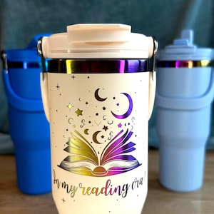 Puede incluir: Vaso t&eacute;rmico blanco roto con tapa y asa. El vaso presenta un dise&ntilde;o de libro abierto con estrellas, lunas y el texto "In my reading era!" en un acabado met&aacute;lico. Se ven otros dos vasos en el fondo.