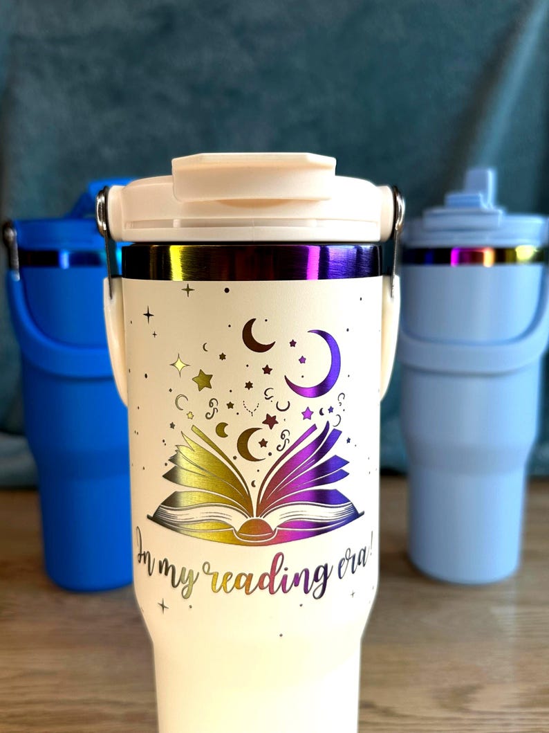 Puede incluir: Vaso t&eacute;rmico blanco roto con tapa y asa. El vaso presenta un dise&ntilde;o de libro abierto con estrellas, lunas y el texto "In my reading era!" en un acabado met&aacute;lico. Se ven otros dos vasos en el fondo.