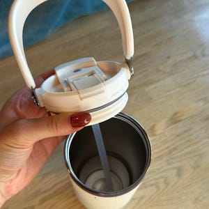 Puede incluir: Vaso t&eacute;rmico aislado de color crema con asa y tapa blancas. La tapa est&aacute; abierta, revelando una pajita en el interior. El vaso tiene un interior de acero inoxidable y un borde plateado. El asa es arqueada y est&aacute; unida a la tapa con bisagras met&aacute;licas.