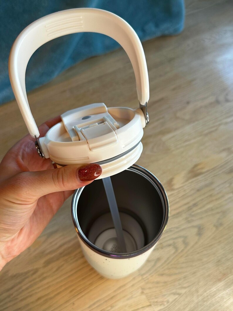 Puede incluir: Vaso t&eacute;rmico aislado de color crema con asa y tapa blancas. La tapa est&aacute; abierta, revelando una pajita en el interior. El vaso tiene un interior de acero inoxidable y un borde plateado. El asa es arqueada y est&aacute; unida a la tapa con bisagras met&aacute;licas.