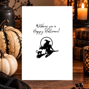 Op de afbeelding: Een witte rechthoekige wenskaart met de zwarte tekst "Witching you a Happy Halloween!" boven een zwarte silhouetafbeelding van een heks met lang haar en een puntmuts op een bezemsteel met een kleine kat voor een volle maan.