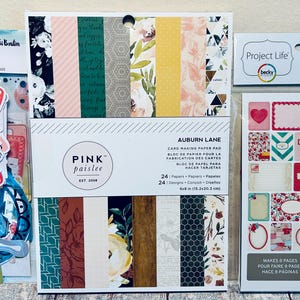 May include: A collection of scrapbooking supplies, including paper pads, stickers, and embellishments. The paper pad features floral and geometric patterns. The sticker sheets include labels and decorative elements. The color palette includes pink, green, and yellow.
