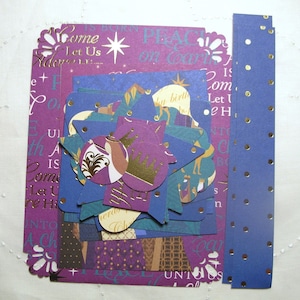 May include: A collection of decorative paper cutouts in shades of purple and blue, featuring gold foil accents, star shapes, and nativity-themed illustrations. The set includes rectangular sheets with text, circular pieces, and two long strips with gold dots.