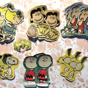 May include: A collection of die-cut paper figures featuring characters from the Peanuts comic strip. The figures include Charlie Brown, Lucy, Sally, and Snoopy, in various poses and outfits. The figures are in yellow, red, and black.