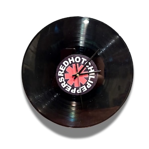 May include: A black vinyl record repurposed as a wall clock. The clock face features the Red Hot Chili Peppers logo in red and white. The clock hands are black and the clock is set against a white background.