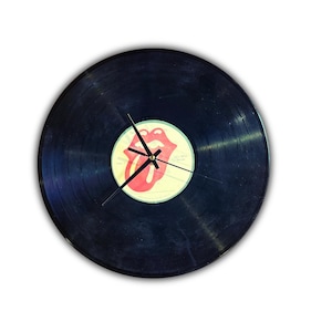 May include: A circular wall clock crafted from a black vinyl record. The center label features the iconic red lips and tongue logo. Three thin black clock hands are mounted in the center, extending across the grooved surface of the record.