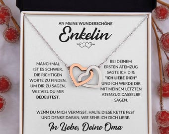 May include: A silver necklace with two interlocking heart pendants, one rose gold and one silver, presented in a gift box. The box contains German text, including the word "Enkelin" and a loving message.