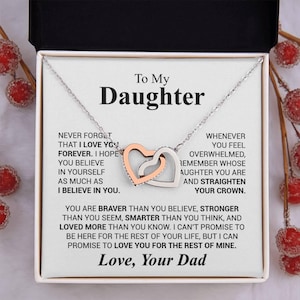 Puede incluir: Un collar plateado con dos colgantes de corazón entrelazados, uno de oro rosa y otro plateado, presentado en una caja de regalo. La caja contiene un mensaje para una hija de su padre, con palabras de amor y ánimo.