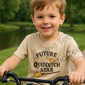 May include: A young child wearing a tan t-shirt with the words "FUTURE QUIDDITCH STAR" in black, along with a golden snitch and a broom graphic. The child is smiling and riding a bicycle.