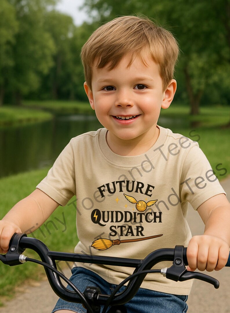 May include: A young child wearing a tan t-shirt with the words "FUTURE QUIDDITCH STAR" in black, along with a golden snitch and a broom graphic. The child is smiling and riding a bicycle.