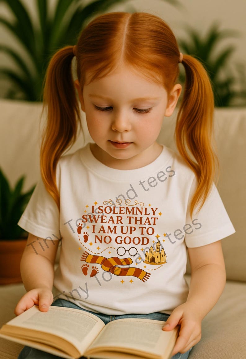 May include: A white t-shirt with the text "I SOLEMNLY SWEAR THAT I AM UP TO NO GOOD" in orange and brown. The shirt features a graphic of a scarf, glasses, footprints, and a castle. The child is reading a book.