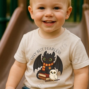 Op de afbeelding: Een lachende peuter draagt een beige t-shirt met een afbeelding van een zwarte draak met een bril en sjaal, en een witte uil. De tekst "TINY BUT FEARLESS" staat boven de draak. Het kind draagt een blauwe spijkerbroek.