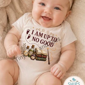 May include: A cream-colored short-sleeve baby bodysuit featuring the text I AM UP TO NO GOOD in dark brown letters. The design includes illustrations of three small glass potion bottles, a stack of two books, and a magic wand with gold sparkles.