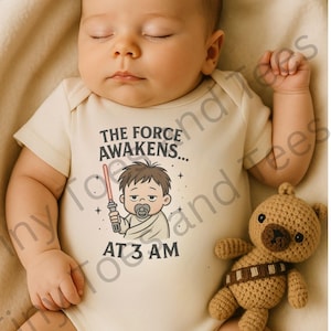 Puede incluir: Un bebé dormido con un body beige con el texto "THE FORCE AWAKENS... AT 3 AM" y una ilustración de dibujos animados. Un pequeño oso de peluche marrón tejido con una bandolera descansa junto al bebé.