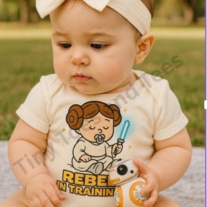 May include: A baby wearing a cream-colored onesie with a cartoon graphic of Princess Leia holding a lightsaber and a pacifier. The text "REBEL IN TRAININ" is below the graphic. The baby is also wearing a cream-colored bow headband and holding a small BB-8 toy.
