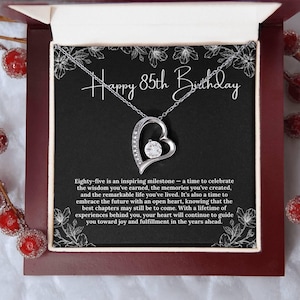 Puede incluir: Collar con colgante plateado en forma de corazón con una piedra preciosa transparente, presentado en una caja roja oscura. El fondo negro tiene el texto "Happy 85th Birthday".