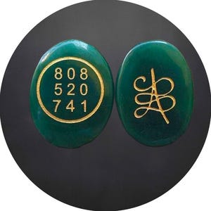 Op de afbeelding: Twee ovale, groene stenen met gouden details. Eén steen heeft de nummers 808, 520 en 741 in een gouden cirkel. De andere heeft een gouden, gestileerd ontwerp. De stenen staan tegen een zwarte achtergrond.