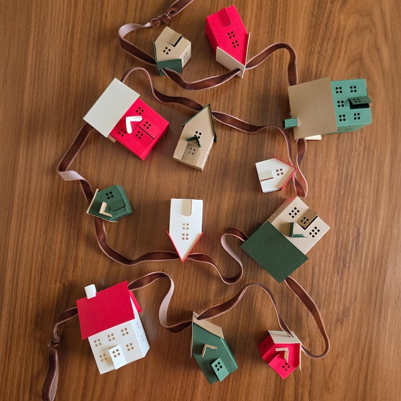 K&ouml;nnte beinhalten: Eine Anordnung von Miniatur-Papierh&auml;usern in verschiedenen Farben, darunter Rot, Gr&uuml;n und Beige, liegt auf einer Holzoberfl&auml;che. Jedes Haus hat kleine Fensterausschnitte und ist durch ein braunes Band verbunden, was eine festliche Dekoration ergibt.
