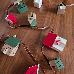 K&ouml;nnte beinhalten: Eine handgefertigte Papierhausgirlande mit H&auml;usern in Rot, Gr&uuml;n und Beige. Die H&auml;user sind auf einem braunen Band aufgereiht und haben kleine quadratische Fenster. Die Girlande wird auf einer Holzoberfl&auml;che pr&auml;sentiert.