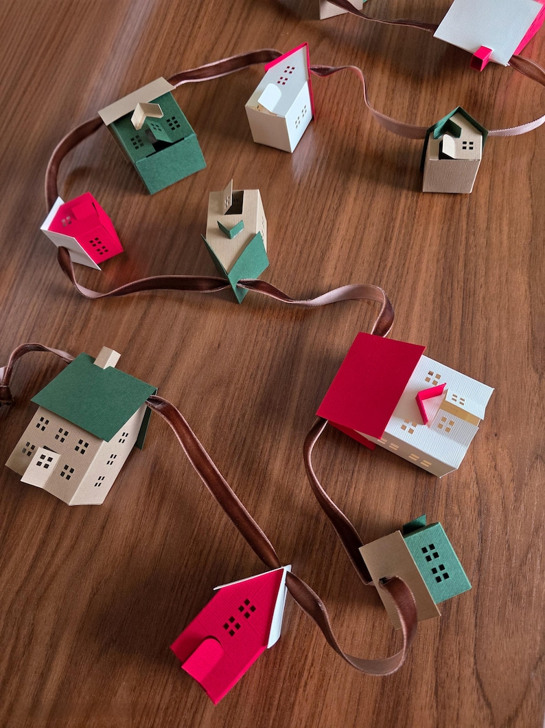 K&ouml;nnte beinhalten: Eine handgefertigte Papierhausgirlande mit H&auml;usern in Rot, Gr&uuml;n und Beige. Die H&auml;user sind auf einem braunen Band aufgereiht und haben kleine quadratische Fenster. Die Girlande wird auf einer Holzoberfl&auml;che pr&auml;sentiert.
