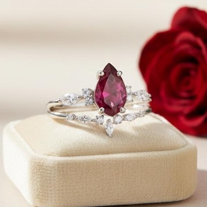 May include: A silver ring featuring a pear-cut, deep red gemstone and smaller white stones. The ring is displayed on a beige velvet box, with a red rose in the background.