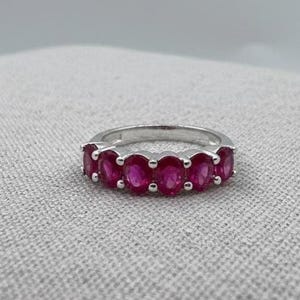 Puede incluir: Un anillo de plata con una fila de piedras preciosas rojas brillantes de corte ovalado. Las piedras están engastadas juntas, creando una banda continua de color. El anillo se muestra sobre una superficie texturizada de color gris claro.
