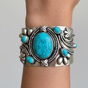 May include: Silver cuff bracelet featuring a large oval turquoise stone at the center, surrounded by smaller turquoise stones and floral designs. The bracelet is wide and has an antique finish, showcasing intricate details.