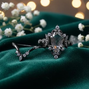 May include: A black engagement ring set with a large, clear, faceted gemstone and smaller accent stones. The ring is paired with a delicate band, both set on a dark green velvet surface, with white flowers and bokeh lights in the background.