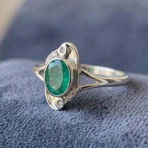 May include: A silver-toned ring featuring a central oval-cut green gemstone. The setting includes a split shank design and is accented with small, clear, round-cut stones embedded in the metal frame surrounding the primary green oval stone.