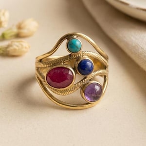 May include: A gold-toned metal ring featuring an open, wavy band design with textured detailing. The ring is set with four distinct gemstones: a small turquoise, a deep blue lapis lazuli, a large oval ruby, and a round purple amethyst stone.