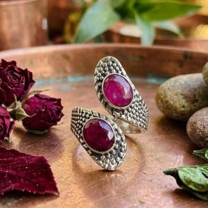 May include: A silver-toned ring with two oval, deep red gemstones. The ring's band features a textured, dotted design. The gemstones are set in a bezel setting. The ring is displayed on a copper-colored surface.
